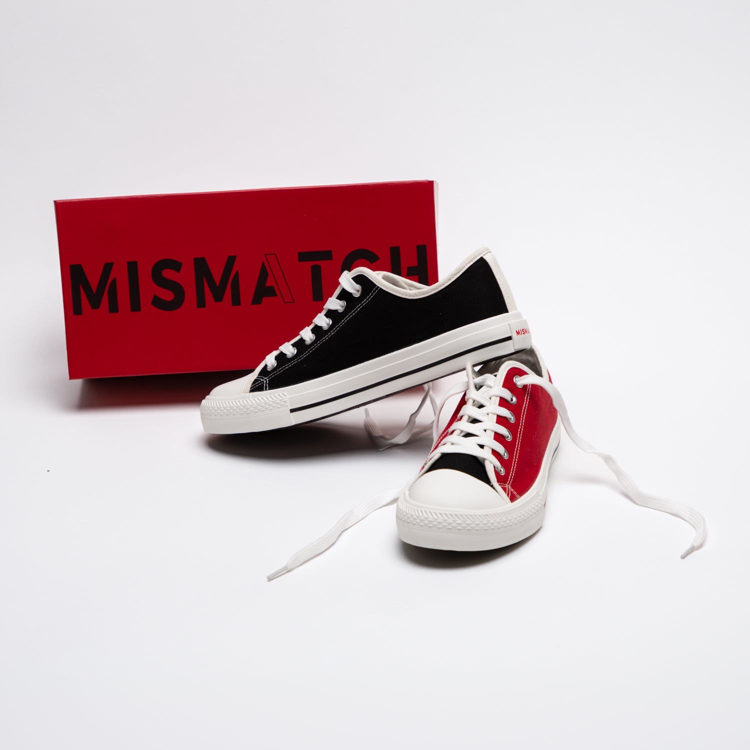 Pair of red and black mismatched sneakers stacked next to Mismatch logo shoe box against white backdrop